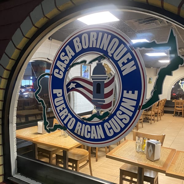 Casa Borinquen restaurant's front window with its logo, showing the interior with tables and chairs.